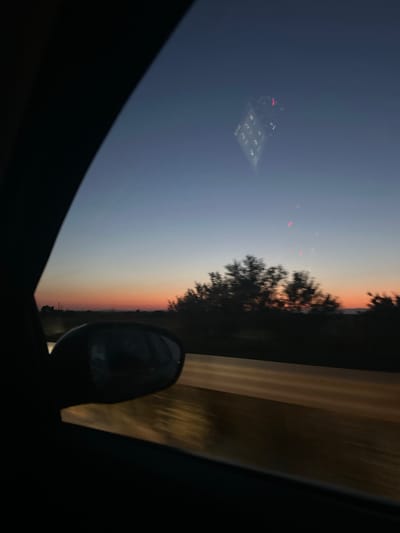 The image depicts a view from inside a vehicle at dusk, looking out the side window towards a twilight sky. A car's side mirror is visible in the foreground, reflecting the road and trees. The sky transitions from a deep blue at the top to warm orange and pink hues near the horizon, indicating sunset. Silhouetted trees line the horizon, with some appearing blurred due to motion. A faint reflection of a building with illuminated windows can be seen in the upper portion of the sky. The road surface, visible through the window, is illuminated by streetlights or headlights, creating a streaky, golden effect. The overall impression is one of a journey through the countryside at the end of the day. There are no people visible in the image. The location is stated to be the city of Skutare, Bulgaria.