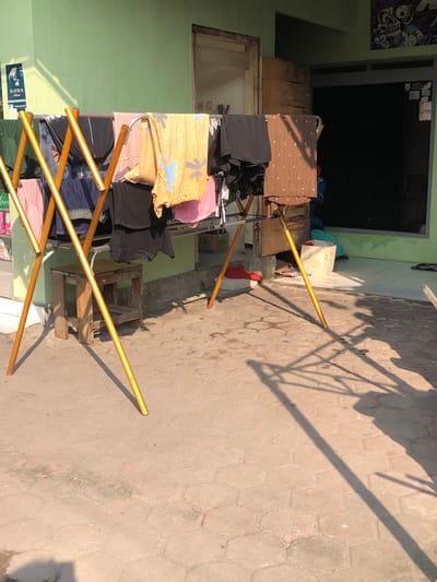 The image shows a clothes drying rack in front of a building in Balonggabus, Indonesia. The drying rack is made of golden metal and is filled with various pieces of clothing, including shirts, pants, and what appears to be a patterned fabric. There is a small wooden stool placed beneath the drying rack. The ground is paved with hexagonal tiles. In the background, the building has a light green wall and a partially open wooden door. Sunlight casts shadows from the drying rack onto the tiled ground, indicating it is daytime.