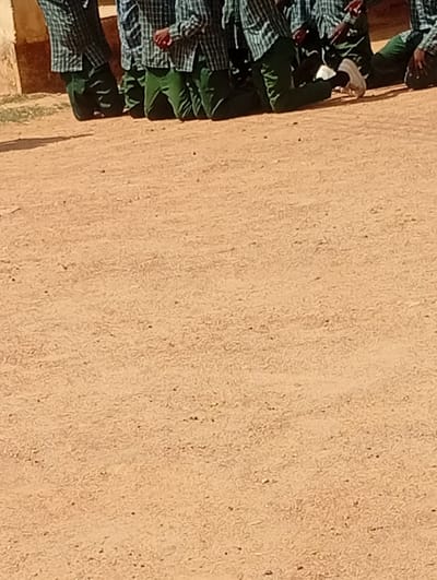The image shows the legs and torsos of several individuals, likely students, kneeling on a sandy or dirt ground. They are wearing plaid shirts and green trousers. One person on the right is wearing white sneakers, while another on the left is wearing black shoes. The background on the left shows a portion of a building, possibly made of mud bricks or concrete. The scene appears to be outdoors, in a dusty or sandy area. The location is identified as Dutse, Nigeria.