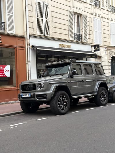 A grey Mercedes-Benz G-Wagen SUV is parked on a paved street next to a multi-story building. The vehicle, visible from the front and side, features large, dark alloy wheels and a distinctive grille. Its French license plate, "GK-806-YM," is clearly visible on the front.
The building displays classic Parisian architectural elements, including light-colored stone or stucco facades, multiple windows with white louvered shutters, and decorative wrought-iron window guards on some lower floors. A black awning extends from a ground-floor shop, prominently displaying "BERECHIT Concept store" in golden lettering. A smaller black sign above the awning also shows "BERECHIT." The building's facade bears the number "11."
Adjacent to the "BERECHIT" store, another ground-floor unit features a wooden-framed storefront. A red and white real estate sign, partially obscured but legible, reads "LOCAL DISPONIBLE," "Orpi VALLOIS," and includes the phone number "01 41 05 55 55." This sign, particularly "VALLOIS," confirms the location context of Levallois-Perret, France.
The street is asphalt with white parking lines. The scene appears to be set during daytime, with soft, diffused lighting suggesting an overcast sky. There are no people visible in the image.