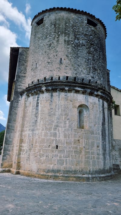 The image shows a close-up, low-angle view of the round tower of the Church of Sant Pere de Montagut, located in Montagut i Oix, Spain. The tower is constructed of rough-hewn stone blocks and features a series of small, dark openings near the top, characteristic of defensive architecture. A decorative stone band with corbels encircles the upper section of the tower, supporting a tiled roof. A narrow, arched window is visible on the lower part of the tower's curved wall. To the left, a portion of an adjacent building with a sloping roof is visible. The ground in front of the tower is paved with irregular stone slabs. The sky, a clear blue with some white clouds, is visible in the background.
