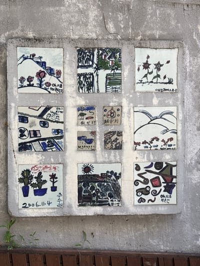 This image depicts a public art installation embedded into a weathered concrete wall in 종로구, South Korea. The central feature is a large, rectangular, slightly raised frame, approximately 1.5 meters in height and 1 meter in width, containing a mosaic of thirteen hand-painted ceramic tiles.
The tiles are arranged in a structured grid: three large square tiles occupy the top row, followed by a middle row comprising one large square tile on the left, a 2x2 cluster of four smaller, irregular square tiles in the center, and one large square tile on the right. The bottom row consists of three large square tiles. Each tile features a unique, colorful, and stylized painting, indicative of a naive or amateur artistic style, likely by children or community members. The subjects depicted on the tiles include landscapes with rolling hills, flowers, and power lines; abstract floral patterns; scenes with houses and bamboo; various abstract shapes and patterns; a stylized urban interior; small animals; rain clouds; birds flying over hills; and potted plants. Several tiles bear inscriptions in Korean, typically including dates (e.g., "2006.1.18," "2006.11.4," "2006.11.8") and names, identifying the presumed artists.
The concrete wall surrounding the tile display exhibits significant signs of wear, including cracking, discoloration, and peeling paint, indicating age and exposure to the elements. Below the tile installation, a section of dark brown wooden slats, possibly a low fence or bench, is visible. Small green plants are seen growing from crevices along the base of the concrete wall and the wooden structure. The scene is illuminated by natural light, casting subtle shadows on the wall's surface and the wooden slats. No individuals are present within the frame.