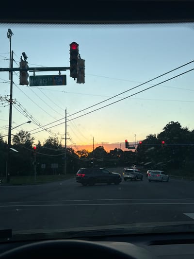 The image appears to be taken from inside a vehicle in Piscataway Township, United States of America. The view is looking towards an intersection with a red traffic light. A street sign indicates "Metlars Ln" and the number "609." Power lines stretch across the sky. There are several cars on the roadway. The sky shows colors suggesting sunset or sunrise. Trees and some streetlights are visible. The driver’s-side dashboard and part of the interior of the car are also in the frame.
