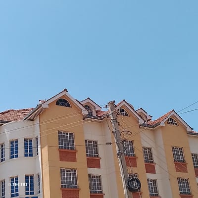 The scene depicts the exterior of a multi-story building and a utility pole, situated in the city of Machakos Central ward, Kenya, under a clear blue sky. The building, which occupies the lower and central portions of the frame, features primarily light yellow and off-white facades. Numerous grid-patterned windows with white or blue frames and dark panes are visible across its various levels. Many light yellow sections incorporate a red-brown brick-patterned fascia directly below the windows. The building is topped with a reddish-brown tiled roof system, characterized by multiple dormer windows, some featuring arched tops. A curved architectural element is present on the left side of the structure. A dark-colored utility pole, likely concrete, stands prominently in the mid-right of the image, partially obscuring the building. Multiple wires are attached to the pole and extend horizontally across the frame. A coiled bundle of dark cables is secured to the lower portion of the pole. No individuals or active interactions are visible. The upper two-thirds of the image displays a clear, bright blue sky, indicating daytime conditions. A "© TECNO" watermark is present on the lower-left facade of the building.