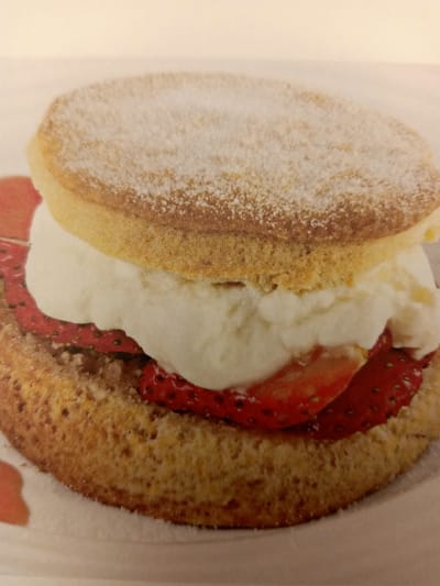 The image shows a close-up of a strawberry shortcake. The cake consists of two golden sponge cake layers filled with whipped cream and sliced strawberries. The top layer of the cake is dusted with powdered sugar. The cake is placed on a white surface, possibly a plate or tablecloth. There are some indistinct red and orange smudges on the white surface around the cake, suggesting some of the strawberry filling or sauce has spilled. The lighting appears to be soft and even. The image is a food photograph, likely for a recipe or advertisement.
The city of Rumaithiya, Kuwait is not depicted or referenced in this image. The image is a purely culinary depiction.