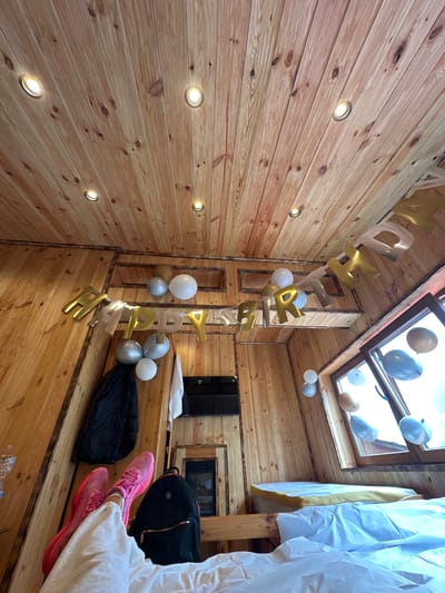 The image shows a cozy, wooden-paneled room, likely a bedroom, decorated for a birthday celebration. A "Happy Birthday" banner made of gold letters is strung across the ceiling. Several balloons in gold, silver, and white are also scattered around the room, some attached to the banner and others floating near the window.

The room appears to be a part of a cabin or a small house, with wooden walls and a wood-paneled ceiling featuring recessed lighting. There's a window with a view of an overcast sky. In the foreground, two pairs of bright pink sneakers are visible, suggesting someone is relaxing in bed. A black backpack rests on the bed next to the feet. In the background, there's a bed with white bedding, a television mounted on the wall, and a fireplace-like structure. There's also a dark jacket hanging on the wall.

The location is identified as Laništa, Montenegro. The overall scene conveys a festive and intimate atmosphere, celebrating a birthday in a rustic setting.