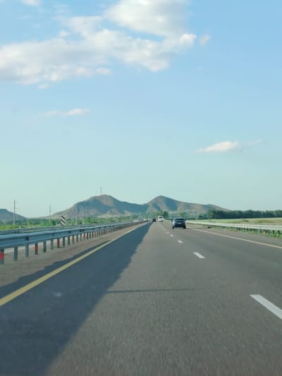 The image depicts a view from a vehicle traveling on a highway towards a mountainous landscape under a partly cloudy sky. The highway has multiple lanes, marked by white lines and a yellow line on the left side. A metal guardrail runs along the left edge of the road, with some warning signs visible. Several cars and a van are driving on the highway in the distance. The foreground consists of the asphalt road surface and its shadow. The middle ground shows the guardrail, fields, and trees. The background features rolling hills and mountains, one of which has a communication tower on its peak. The overall scene suggests a sunny day with clear visibility, likely on a road in or near the city of Chkalovo, Kazakhstan, given the mountainous terrain and vegetation.