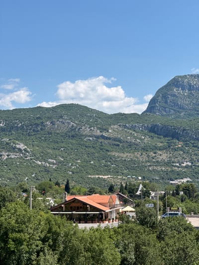 In the city of Cerovo, Montenegro, this image captures a rural landscape with a focus on a building resembling a restaurant or cafe, situated amidst lush greenery. The building features a prominent orange tiled roof and large glass windows, suggesting an establishment open to the outdoors or with scenic views. In front of the building, potted plants line the pavement, and a yellow canopy is erected. Beyond the building, the terrain rises into heavily wooded hills and mountains, characteristic of Montenegro's topography. The sky above is a clear blue with scattered white clouds. A few vehicles are visible, including a white van and a darker car parked on a road near the building, indicating a degree of accessibility. The overall impression is one of a tranquil, natural setting with a touch of civilization.