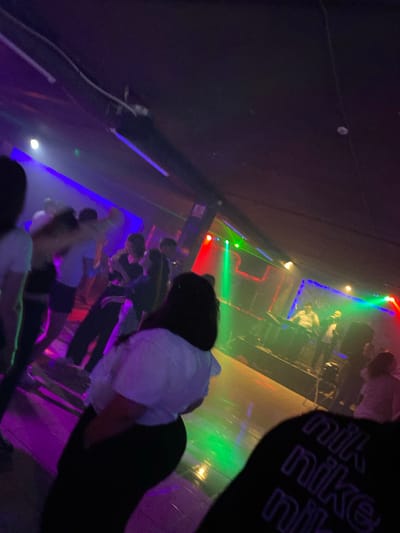The image depicts a dimly lit indoor venue, possibly a nightclub or concert hall, in Nisporeni, Moldova. The scene is characterized by vibrant, colored lighting, primarily blues, purples, and greens, casting dynamic beams across the space and illuminating the floor. A group of people are visible, some dancing and socializing, while others are gathered on or near a stage area. On the stage, two individuals appear to be performing or operating equipment, illuminated by spotlights. The atmosphere suggests a lively event, with the lighting and the presence of a crowd contributing to a festive or energetic ambiance. The overall image quality is somewhat blurry, consistent with a low-light environment and movement within the frame.