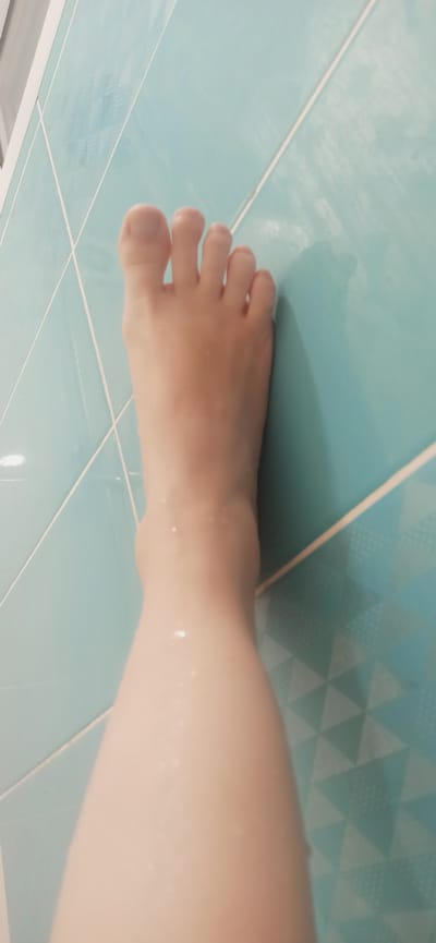 The image shows a close-up view of a person's left foot and lower leg against a background of light blue tiles. The tiles are arranged in a grid pattern, with white grout lines. The foot and leg appear wet, with small droplets of water visible on the skin. The lighting suggests the photo was taken indoors, possibly in a bathroom. The image does not contain any elements that would suggest a specific location within the city of Asbest, Russia.