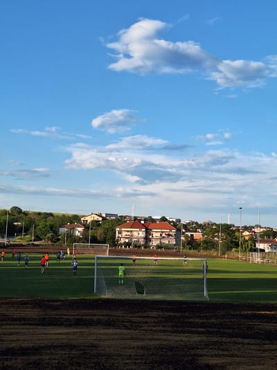 An outdoor football pitch in Ezerovo, Bulgaria, is observed under a bright blue sky with scattered white cumulus clouds, suggesting a late afternoon or early evening setting due to the soft, warm light. In the immediate foreground, a strip of dark, disturbed earth with visible indentations occupies the lower portion of the frame. A white football goal with a net stands prominently on the right side of the midground. On the green grass pitch, multiple individuals are engaged in a football game or practice. One player in a bright green uniform is positioned within the goal area, facing away from the camera. Other players are dressed in blue and orange/red jerseys, with one blue-jerseyed player featuring the number "19" on his back. A second white goal is visible at the far end of the pitch. Beyond the playing field, a dense line of green trees and bushes separates the pitch from a residential area. Several multi-story houses with light-colored facades and red-tiled roofs are scattered across the background. Further back, a white cellular or communication tower rises above the distant structures. Utility poles and power lines are visible, particularly on the left side of the background, alongside what appears to be a road.