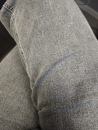 A close-up, top-down shot shows a pair of dark grey denim jeans. The fabric is textured with the characteristic weave of denim. A subtle crease runs diagonally across the right side of the frame, suggesting the material has been folded or worn. The stitching along a seam is visible on the left side, running vertically. The lighting creates some highlights and shadows, emphasizing the texture of the fabric. The background is dark and out of focus. This image is a detail of clothing, not a scene that can be located in a specific city or environment.