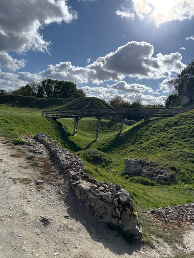 The image captures a vibrant outdoor landscape featuring undulating green terrain, likely earthworks or mounds, under a bright, partly cloudy sky. The scene is set in what appears to be a historical or natural park in King's Lynn and West Norfolk, United Kingdom.
In the mid-ground, a wooden footbridge or walkway with railings spans across a dip in the grassy mounds, connecting two higher points. The slopes of the mounds are covered in lush green grass, illuminated by the sun, creating varied shades and shadows. Scattered trees are visible in the distance and nestled among the mounds, adding to the natural environment.
In the immediate foreground and extending along the right side of the image, there is a rough stone wall or the remnants of a structure, suggesting ancient ruins or a boundary. Adjacent to this wall is a dirt or gravel path, hinting at pedestrian access.
No people are visible, but a small, dark bird can be seen soaring against a large cloud in the sky.
The time of day is bright, likely late morning or early afternoon, with the sun shining intensely from the upper left, casting strong shadows from the wooden bridge and terrain features. The weather is clear and fair, characterized by a blue sky dotted with large, puffy white cumulus clouds, creating a dynamic backdrop. The overall impression is one of tranquility and natural beauty, characteristic of a historical or nature preserve. There is no visible text in the image.