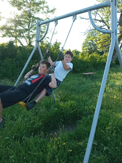 Two young boys are on a swing set in a grassy area. The boy in front is wearing a black t-shirt with a graphic print, black shorts, and black shoes. He is holding onto the swing chains with gloved hands. The boy behind him is wearing a white t-shirt and patterned shorts, also holding onto the swing chains with gloved hands. The swing set is made of metal with grey poles. The background consists of trees and bushes. In the distance, there is a small wooden table with red legs. The sun is setting, casting a warm light on the scene. There is no information about the specific location in the city of Zuya, Ukraine.