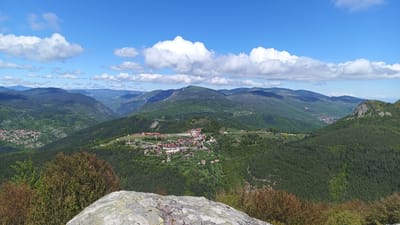 The image shows a panoramic view of a mountainous landscape under a blue sky with scattered white clouds. In the foreground, there is a rocky outcrop with some sparse vegetation. A village with red-roofed buildings lies in a valley between the mountains. The scene is located in or around Sini Vrah, Bulgaria. There are no people visible in the image. The environment is primarily mountainous with green trees and vegetation.