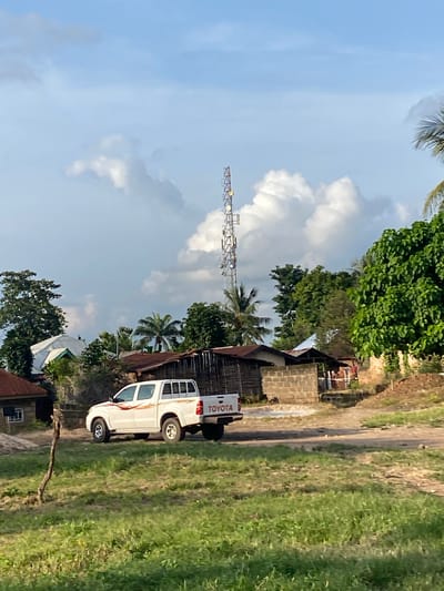In Nguzu Edda, Nigeria, under a partly cloudy sky, a white Toyota pickup truck is parked on a dirt road. The truck has distinctive red and orange pinstripes along its side and "TOYOTA" is visible on the tailgate. To the left of the truck, several buildings with corrugated metal roofs and brick walls are visible. A tall communication tower with multiple antennas stands in the background, silhouetted against the sky. Lush green trees and palm trees surround the structures, and the ground is covered in dry grass and some patchy green vegetation. The overall impression is a rural or semi-urban setting with signs of development and infrastructure.