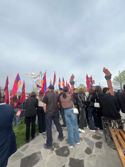 A scene in Akhuryan, Armenia, presents a gathering of people near a series of Armenian flag arrangements. There are stone sculptures and decorative elements present. Individuals, some holding the flags, are standing or positioned along a paved stone path. The atmosphere suggests an event or commemoration. The sky is overcast.