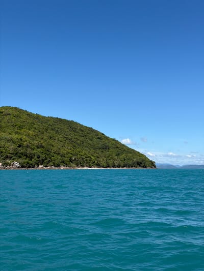 A vast, clear blue sky dominates the upper portion of the image. Below the sky, a lush, green, tree-covered hillside slopes down from the upper left to the middle right of the frame. The hillside meets a narrow strip of sandy beach. The foreground is filled with the tranquil, turquoise waters of the sea, showing gentle ripples and a few small whitecaps. The overall scene suggests a tropical island landscape, likely in Baan Thong Krut, Thailand, under a bright, sunny day. There are no people or animals visible in the image.