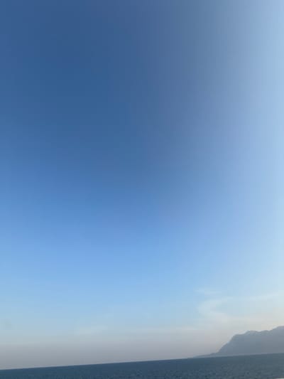 The scene depicts a vast expanse of clear sky over a body of water, located in Kato Karioutes, Greece. The upper approximately 80% of the image is dominated by a clear blue sky, gradually lightening in hue towards the horizon. The lower approximately 15-20% of the image features a dark blue body of water, exhibiting minor surface ripples. In the far distance on the right side, a light-colored, hazy landmass, likely a mountain range or coastline, extends along the horizon line. No people, subjects, objects (other than natural elements), actions, or interactions are visible within the frame.