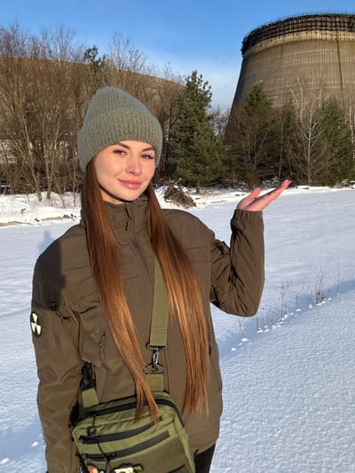 A young woman stands outdoors in a snowy winter landscape, with a prominent derelict cooling tower in the background, strongly suggesting a location like Pripyat, Ukraine, reinforced by the radioactive symbol patch on her jacket. She has long brown hair and is dressed in winter attire: a grey knit beanie, an olive green puffer jacket, and a matching olive green shoulder bag. She smiles towards the camera, her right hand raised in a wave.

The setting is bright and sunny with a clear blue sky. Bare deciduous trees and evergreen pines dot the snow-covered ground. The large, concrete, derelict cooling tower dominates the left background, appearing weathered. A yellow circular patch with a black radiation hazard symbol is visible on her left jacket sleeve. The ground is uniformly covered in fresh white snow. No explicit text is visible in the image beyond the symbol.