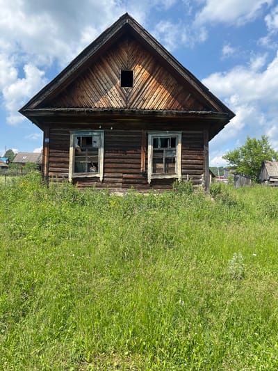 An old, weathered wooden house stands in a field of tall green grass under a blue sky with scattered clouds in Новомуллакаево, Russia. The house has a steeply pitched roof covered in wooden shingles. Two windows on the front of the house are broken, with glass shards visible. A small dark square opening is present in the upper part of the gable. The logs of the house are dark brown and appear aged. A blue sign with the number "49" is affixed to the left side of the house. In the background, other structures and trees are visible, suggesting a rural or semi-rural setting. The grass is overgrown and dense, reaching up to the bottom of the windows.