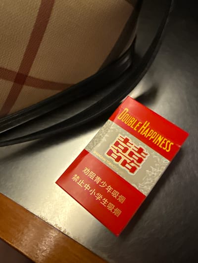 The image shows a red cigarette pack with "DOUBLE HAPPINESS" written in yellow text. The pack also features Chinese characters and a stylized design of the Chinese character "囍" (shuāngxǐ), which symbolizes double happiness and is traditionally used for weddings. Below the "囍" symbol, it reads "SHANGHAI TOBACCO". The pack is placed on a surface that appears to be a table or desk, with a portion of a checkered fabric and a dark, curved object, possibly part of a bag or chair, visible in the foreground. In the background, there is a dark, textured surface, likely a tabletop. The lighting suggests it might be indoors, with some ambient light creating shadows. The image's composition and the presence of the cigarette pack imply an everyday scene. The Chinese text on the pack translates to "Discourage adolescent smoking" and "Forbid junior and senior high school students to smoke," indicating a public health warning. The context provided places this scene in Jing'an District, China.