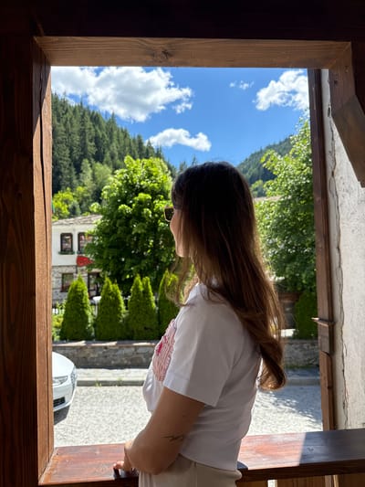 A medium shot shows a woman with long brown hair, wearing sunglasses and a white t-shirt, standing and looking out from a wooden window frame. The background reveals a scenic view of Shiroka Laka, Bulgaria, with a cluster of traditional white houses nestled among lush green trees and a steep, forested mountainside under a bright blue sky with scattered white clouds. In the foreground, a white car is partially visible parked on a cobblestone street. The woman has a tattoo on her left forearm.