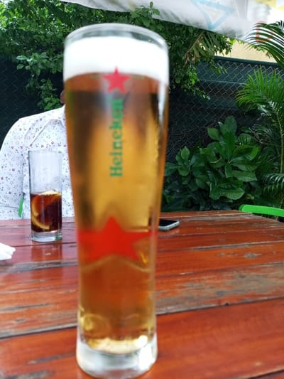 A glass of Heineken beer sits on a wooden table, a portion of which is visible, at a restaurant or bar. The glass is half full. A person's arm and part of a white, patterned shirt can be seen in the background. A clear glass with a dark liquid and a slice of lemon or lime in it is visible on the table to the left of the beer glass. A dark, cellular phone is lying on the table to the right. Lush greenery is visible in the background, and a green chair can be glimpsed in the upper right corner.  This image is likely taken at a bar or restaurant in Maputo, Mozambique, at the coordinates -25.976 latitude and 32.591 longitude.