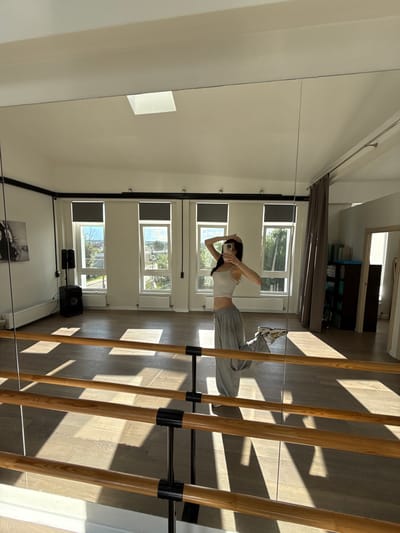 The image captures a person taking a selfie in a dance studio. The setting appears to be a spacious room with wooden floors and a large mirror covering one wall. A barre, made of multiple wooden poles, runs along the right side of the frame, casting distinct shadows from the sunlight streaming through the windows.

The subject, seen in the reflection of the mirror, is a person with dark hair, wearing a white tank top and grey sweatpants. They are holding a smartphone and appear to be adjusting their hair. Their posture suggests a relaxed, casual pose.

Several windows are visible in the background, revealing a view of trees and buildings, indicating an outdoor scene. The lighting suggests it is daytime, with strong sunlight creating harsh shadows. Some radiators are visible beneath the windows, and a speaker system and a black cabinet are placed on the floor. A piece of artwork, possibly a black and white photograph, hangs on the wall.

The overall impression is one of a casual moment captured within a functional space, possibly during a dance practice or break. The presence of the barre and the mirror strongly suggest a dance or movement studio. The sunlight creates a dynamic interplay of light and shadow, adding a sense of depth and atmosphere to the scene. No visible text is present in the image.