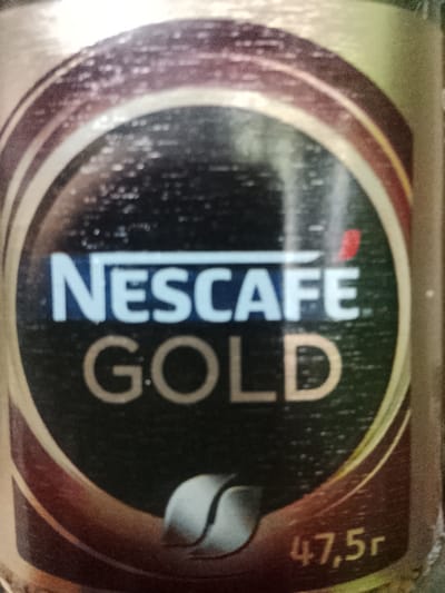 The image shows a close-up of a jar of Nescafé Gold instant coffee. The label is predominantly gold and dark brown with the Nescafé Gold logo in white and red. The text "NESCAFÉ GOLD" is clearly visible. To the bottom right of the label, "47,5г" is written, indicating the weight of the product in grams. There is also a graphic of two stylized leaves. The lighting in the image is somewhat harsh, creating reflections and obscuring some details on the jar. The background is out of focus and appears to be a general indoor environment, but no specific details about Obizarang, Uzbekistan, are visible or implied.
