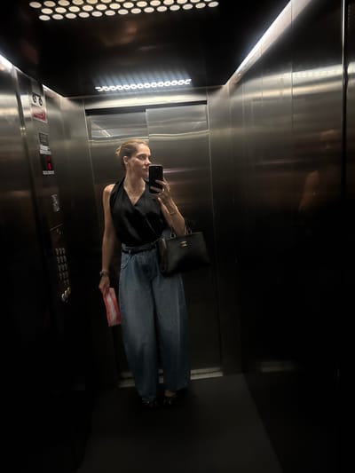 A woman is standing in an elevator, taking a selfie with her phone. She is wearing a black sleeveless top, blue wide-leg jeans, and black patterned shoes. She is holding a red and white object in her left hand and a black handbag with a gold logo in her right hand. The elevator has stainless steel walls and a control panel with buttons and a digital display. The lighting in the elevator is dim, with some reflections on the walls. The image does not provide any information that would allow for the localization of this scene in Loures, Portugal.