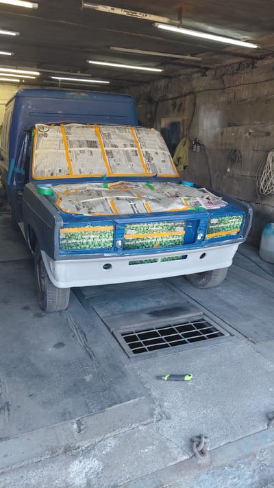 In a dimly lit garage in Belovo, Russia, a blue van is being prepped for painting. The hood and windshield are covered with newspaper and yellow masking tape, suggesting the vehicle is undergoing bodywork or a paint job. The front grille area has been painted a vibrant green with some lettering visible, while the lower bumper section is covered in white primer. The garage environment is characterized by concrete floors, exposed brick walls, and fluorescent lighting. Tools, including a utility knife and chains, are scattered around the floor, hinting at ongoing repair work. A drain grate is visible in the floor, and a blue plastic container is situated to the right. The scene depicts a typical automotive repair setting.