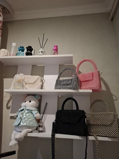 The image depicts an interior scene, likely a room in a private residence, showcasing a collection of handbags and decorative items displayed on white shelves. The shelves are mounted on a textured, light-colored wall.

On the top shelf, from left to right, are a white decorative object, a clear bottle with a white cap, a small blue bear-shaped bottle with "MOSCHINO" visible, a small black bear-shaped bottle, a white diffuser bottle with black reeds, and a pink bear-shaped bottle.

The middle shelf features three beaded handbags. On the left is a white beaded handbag with a perfume bottle on it. To its right is a silver beaded handbag. On the far right of this shelf is a pink beaded handbag.

The bottom shelf displays a plush toy cat dressed in a light blue quilted jacket sitting on the left. Next to the cat is another diffuser bottle. To the right of the cat and diffuser are two more beaded handbags: a black one on the left and a silver one on the right. The handbags are of various sizes and styles, predominantly made of small beads.

The overall impression is one of organized display, with the items carefully arranged on the shelves. There is no indication of the location being specifically in Korenovsk, Russia, or any other specific city. The scene is static and shows objects in a state of rest.