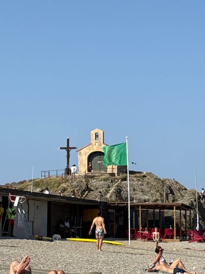 This image depicts a sun-drenched scene on what appears to be a rocky coast, possibly a beach or a waterfront area, with a clear blue sky indicating a pleasant, likely warm day. The orientation suggests it might have been taken during the late morning or early afternoon.

In the mid-ground, a small, light-colored building with arched openings and a pitched roof is visible, perched on the rocks. A green flag flies from a pole in front of this building. To the left of the building, a large wooden cross stands prominently. The surrounding area is a mix of rough rock formations and a paved or cobbled walkway.

Scattered throughout the scene are several people, mostly dressed in swimwear. Some are relaxing, sunbathing, or sitting, while others appear to be walking or standing. One man in patterned swim shorts is captured in a dynamic pose, seemingly mid-stride or stretching, on the walkway near the building. Another individual is lying on their back further down the walkway, with their legs and feet visible in the foreground. Further back, near the building, other people can be seen gathered, some sitting at tables, suggesting the presence of an establishment like a cafe or restaurant. There are also red chairs and tables visible under a wooden structure.

On the far right, more people are seen relaxing on the ground, with their feet prominently in the frame. The overall atmosphere conveyed is one of leisure and recreation, typical of a summer day at a coastal destination.

There is a sign with yellow lettering visible on the building, but the text is too blurry to be read. The location context provided, Collioure, France, suggests a European coastal setting.