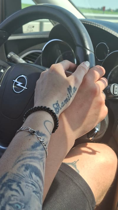 Two people are in the front seats of a car. One person's hands are on the steering wheel, and the other person's hands are resting on the first person's hands. The first person has a tattoo on their hand that says "Kamilla". The first person is also wearing a black beaded bracelet and a silver chain bracelet. The second person is wearing a ring on their finger. The car appears to be driving on a road with a grassy area and trees in the background. The image is taken from inside the car, looking forward. The location is identified as Самахвалавіцкі сельскі Савет, Belarus.