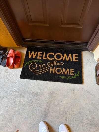 The image shows a doormat with the words "WELCOME TO OUR HOME" in gold lettering on a black background, placed in front of a dark brown door. To the left of the doormat are two orange slip-on shoes and a pair of gray flip-flops. To the right of the doormat, a brown flip-flop is partially visible. In the lower portion of the image, the toes of a person wearing white sneakers are visible. The doormat and shoes are on a concrete surface. The background consists of a textured wall, a door, and the concrete floor. The scene appears to be an entryway to a residential building.