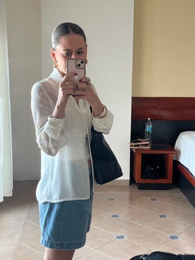 A female individual is depicted in a mirror selfie taken within an indoor setting in Chamilpa, Mexico. She is observed from the waist up, holding a light-colored smartphone with both hands to capture the image. Her dark hair is pulled back, and she wears gold-colored hoop earrings. Her attire consists of a translucent white long-sleeved button-up shirt over a lighter top, paired with light blue denim shorts or a skirt. She has red nail polish, multiple rings on her left hand, and gold-colored bracelets on her right wrist. A black shoulder bag hangs from her right shoulder. The reflected environment shows a room with light beige walls and a tiled floor composed of light brown squares with small blue decorative inserts. A dark brown headboard and a matching nightstand are visible to the right. On the nightstand, a clear plastic water bottle with a blue cap and a pair of light-colored flip-flops are present. A white bed sheet is partially visible adjacent to the nightstand.