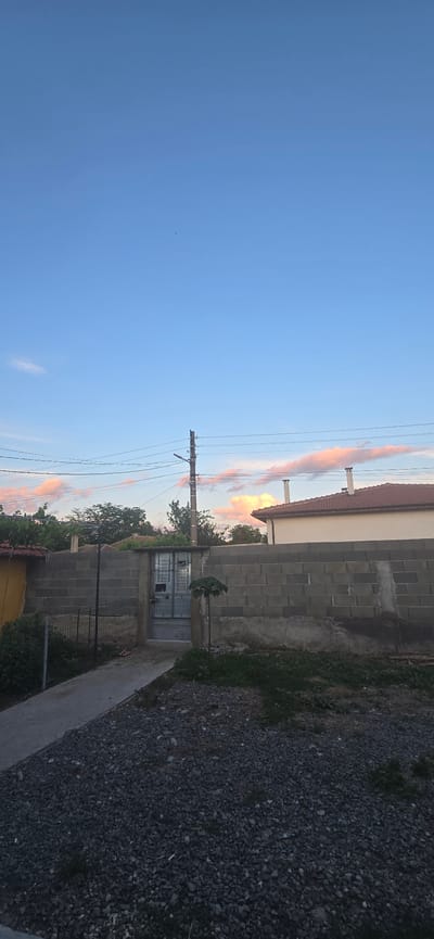 The image depicts an exterior view of a property in Kalchevo, Bulgaria, at what appears to be dusk or early morning given the lighting and the soft hues in the sky. The scene is dominated by a concrete block wall that spans across the middle ground, with a metal gate set into it. To the left of the wall, a black metal fence is visible. Beyond the wall, parts of a building with a red tiled roof and chimneys can be seen, as well as lush green trees. In the foreground, a gravel pathway leads towards the gate, with a paved walkway to its left. Overhead, power lines crisscross the sky, supported by a utility pole with a lamppost attached. The sky itself is a gradient of light blue with scattered wispy clouds tinged with pink and orange, suggesting the presence of the sun near the horizon. The overall impression is of a residential property in a rural or semi-rural setting in Bulgaria. There are no people visible in the image.