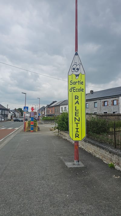 A street sign shaped like a pencil with a cartoon face is positioned on the right side of a paved road. The sign is yellow and displays the text "Sortie d'Ecole" and "RALENTIR" in black lettering, indicating "School Exit" and "Slow Down." The pencil has large, circular eyes, an open mouth with a smile, and is topped by a red pole. To the left of the road, a pedestrian crossing is visible. Further down the road, houses with stone facades and tiled roofs line the street. A black metal fence runs along the right side of the scene, separating the sidewalk from the properties behind it. The sky is overcast, with a cloudy, gray appearance. The overall environment suggests a residential area in a town.