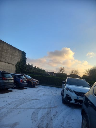 This image depicts a parking area in Noyelles-sous-Lens, France, covered in a thin, uneven layer of snow or frost. The ground displays numerous tire tracks in the snow.
In the left background, a rough-textured concrete wall, partially obscured by climbing ivy, extends upwards. Adjacent to this wall, running across the mid-ground, is a well-maintained green hedge, beyond which distant residential rooftops and bare deciduous trees are visible against the horizon. A flock of small dark birds is visible in flight above the hedge on the left side.
Several vehicles are parked in the lot. From left to right:
* A dark grey SUV, its rear facing the viewer, with a French license plate "SS-425-BS".
* A dark blue compact car, its rear facing the viewer, with a partially visible French license plate beginning "FZ-255".
* An orange SUV, positioned behind the blue compact car, its rear-right quarter visible.
* A white Peugeot 3008 SUV, parked angled towards the viewer on the right, displaying a French license plate "EX-233-XJ".
* The front-right corner of a dark-colored vehicle is partially visible in the extreme foreground on the right.
The sky occupies the upper half of the image, presenting a clear blue hue that transitions to a warmer, yellowish-orange tone near the horizon on the right, indicating either sunrise or sunset. A cluster of illuminated, white and orange-tinged cumulus clouds is present in the upper right quadrant of the sky. No individuals are present in the scene, and no overt actions or interactions are depicted.