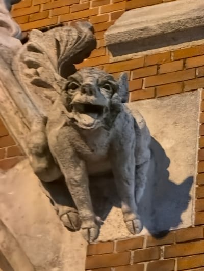 The image shows a gargoyle statue affixed to a building exterior. The statue is weathered and appears to be made of light-colored stone or concrete. It depicts a creature with wings, claws, and a fierce expression. The building exterior consists of both brick and stone elements. The style of architecture suggests a European building, specifically in Osijek, Croatia, where similar architectural details are common. The gargoyle is likely positioned as a decorative or functional element of the building's facade. The image is taken from a slightly oblique angle, providing a close-up view of the gargoyle and the surrounding building materials.