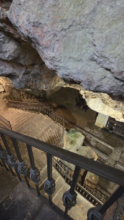 Captured from above, the image depicts a stone staircase descending into a cave in Altura, Spain. The cave entrance, framed by rugged rock formations on the left, leads to a stone-paved area with a white structure and dark objects. Metal railings line the staircase. The perspective emphasizes the depth and verticality of the cave system.