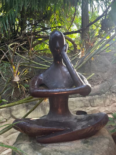 The image shows a dark brown, smooth sculpture of a person in a seated meditation pose, resting on a large, grey, rounded rock. The sculpture is in a lush, green outdoor environment, with dense foliage and trees forming the background. Sunlight filters through the leaves, creating dappled light and shadows. The sculpture appears to be wet, with visible water droplets on its surface. The pose of the sculpture is contemplative, with one arm bent and resting against the head. The setting suggests a natural park, garden, or a similar outdoor recreational area within Khlong Thom Nuea, Thailand.