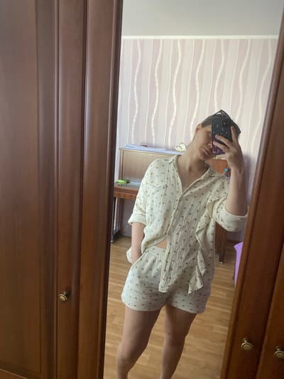 The image shows a young woman in a cream-colored pajama set with a heart pattern, taking a selfie in a mirror. She is wearing a short-sleeved button-down shirt and matching shorts. Her hair is pulled back, and she has sunglasses on top of her head. She is standing in what appears to be a bedroom. To the left of the woman, a dark wooden wardrobe with vertical panels and two round knobs is visible. To the right of the woman, a reflection shows a room with light pink and white striped wallpaper. In the background of the reflection, a wooden upright piano is partially visible, along with a tall purple object and a bottle of water. The floor in the reflection appears to be light wood.

There is no information in the image to suggest the location is the city of Kornilovo, Russia.