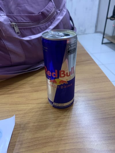 An open 250ml Red Bull Energy Drink can, silver and blue with visible condensation droplets, rests on a light brown wooden table. The can displays the "Red Bull ENERGY DRINK" logo and a Portuguese warning label indicating "ALTO EM AÇÚCAR ADICIONADO" (High in Added Sugar). To the left and behind the can is a light purple fabric bag with a metallic zipper. A small, crumpled piece of white paper with blue markings is visible in the lower-left corner of the frame. The background consists of a light-colored wall or tiled floor and a blurred, dark shelving unit. This scene is located within the city of Lauro De Freitas, Brazil.