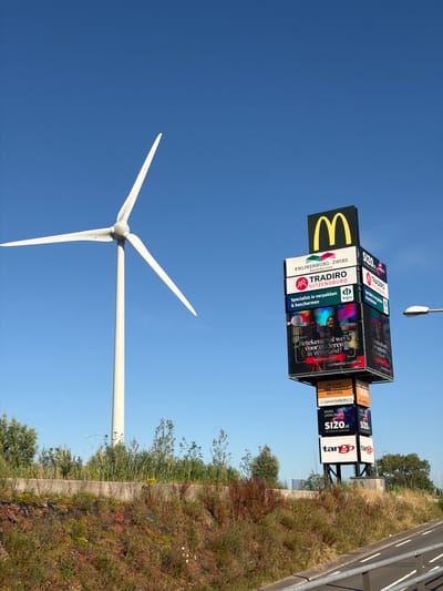 In Maasdijk, Netherlands, a modern wind turbine stands tall against a clear blue sky. To the right of the turbine, a tall, multi-faceted advertising billboard features the iconic McDonald's logo at its peak. The billboard displays various advertisements, including "KNUIJENBURG-ZWIRS KASSENSLOOP," "TRADIRD UITZENDBURO," and "SIGNS LEDSCREENS SIZO.nl," among others. The lower sections of the billboard showcase images of people and text, such as "Betekenisvol werk voor ouderen in Westland?" and "Inleven realiseren beleven." The billboard is mounted on a black metal structure. Below the billboard, the brands "VAN MIERLO" and "tango" are prominently advertised. The foreground is a grassy embankment with various types of vegetation and a concrete barrier, suggesting the billboard and turbine are situated near a roadway. A street lamp is visible in the distance.