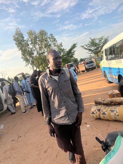 The image captures an outdoor scene in the city of Gbaza, Nigeria, featuring an unpaved, reddish-brown sandy ground under a clear blue sky with scattered white clouds. The strong sunlight casts distinct shadows, suggesting bright daylight.

In the immediate foreground, a dark-skinned man with a beard and short hair, wearing a striped long-sleeved shirt, dark trousers, and dark sandals, stands angled slightly towards the right. He carries a light-colored backpack strap on his right shoulder and holds a smartphone in his left hand, gazing towards the viewer's left with a pensive expression. To his right, several cylindrical rolls of material, marked "ROYAL FOAM" with a geometric pattern, are stacked on the ground alongside a cardboard box labeled "MIMEE Chinchin," a black plastic bag, and a partially visible woven sack.

In the midground to the left, a white SUV or minivan with an open door is surrounded by several men. One man in a light grey garment leans on the vehicle, while another wearing a black backpack examines a smartphone. Nearby, a man in light blue traditional attire stands, potentially holding a striped bag, next to a man in a black t-shirt. To the right midground, a blue and white passenger bus, with "PLATIAU RIDERS" visible on its side, is parked. A person in a light-colored traditional garment stands near the bus.

The background reveals additional vehicles, including a silver sedan, and several indistinct figures along the unpaved road. Multiple trees with green foliage are interspersed throughout the scene, and a low elevation or distant hills are visible on the horizon.