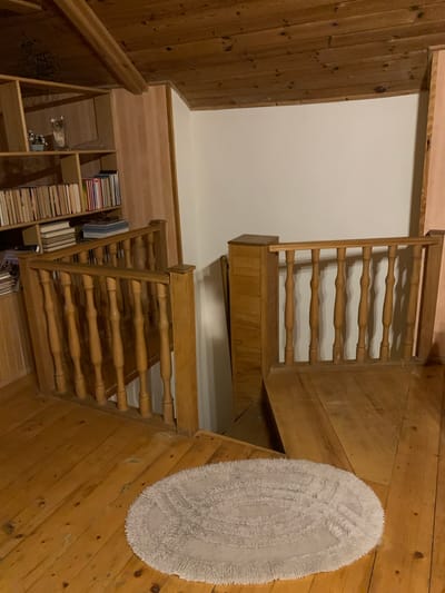 The image shows an interior scene, likely a room with wooden floors and a wooden ceiling with exposed beams. There is a wooden staircase with a wooden railing. A portion of a bookshelf filled with books is visible on the left side of the image, along with some decorative items. A light-colored oval rug is placed on the wooden floor in the foreground. The environment appears to be a residential space, with natural light sources implied by shadows. The city of Mepiskalaki, Georgia is not explicitly depicted or suggested by any elements within the image. There are no people visible in the scene.