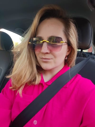 A woman with blonde hair and sunglasses is in the driver's seat of a car. She is wearing a bright pink shirt and a black seatbelt. The sunglasses have pink lenses and gold frames. The woman is looking at the camera. The interior of the car is visible, as well as a reflection of trees and a road on the sunglasses. The image was taken outdoors in the city of Pesyanka, Russia.