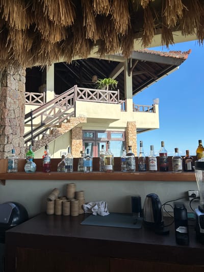 The image captures a bar setup in Senggigi, Indonesia, against the backdrop of a multi-level building under a clear sky.
On the right side, the bar area displays a shelf with various alcohol bottles arranged in a row. Adjacent to the bottles, there are rolls of woven material and a coffee maker. Electrical sockets and equipment cords are visible on the wall behind the bar.
In the background, a building constructed with stone and pale yellow plaster features a wooden staircase with a cross-pattern railing, climbing along the exterior. A thatched roof structure stands to the left of the building, with its straw-like material filling the left foreground. The building's architecture suggests a tropical resort or restaurant setting. The sunlight indicates a daytime scene.